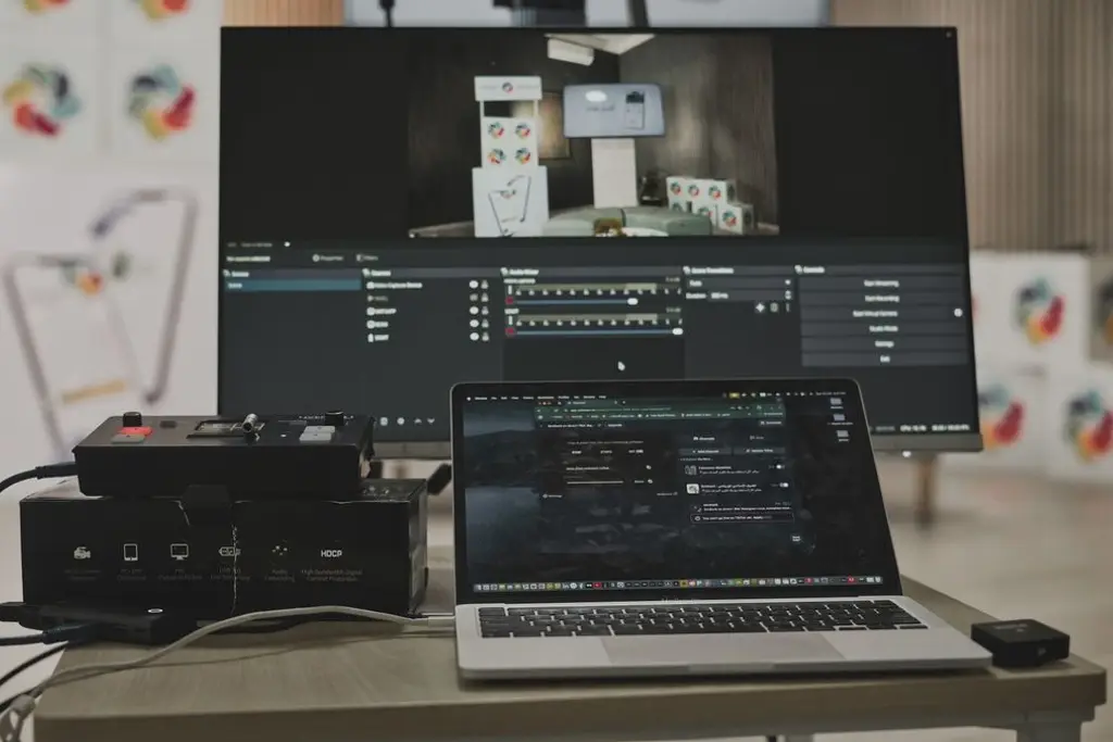 A laptop and monitor showing video editing software on a desk with capture equipment