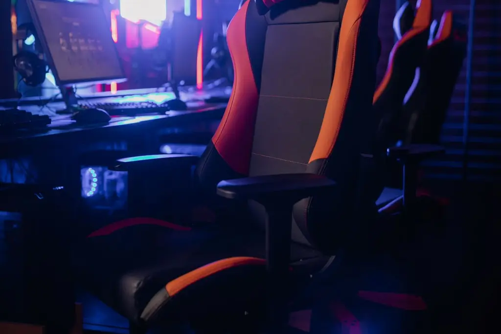 A black and orange gaming chair in front of a monitor in a blue-lit room
